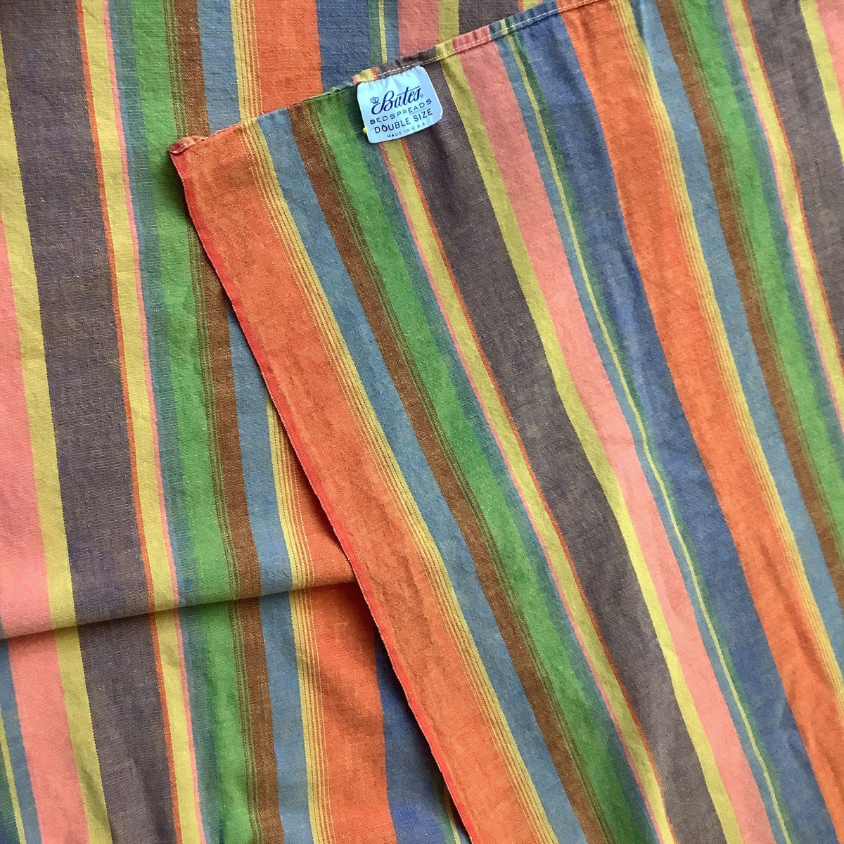 A folded striped Bates vintage bedspread, featuring vertical stripes in orange, green, blue, yellow, and brown. A small white brand tag reading 'Bates' is visible near the top fold.