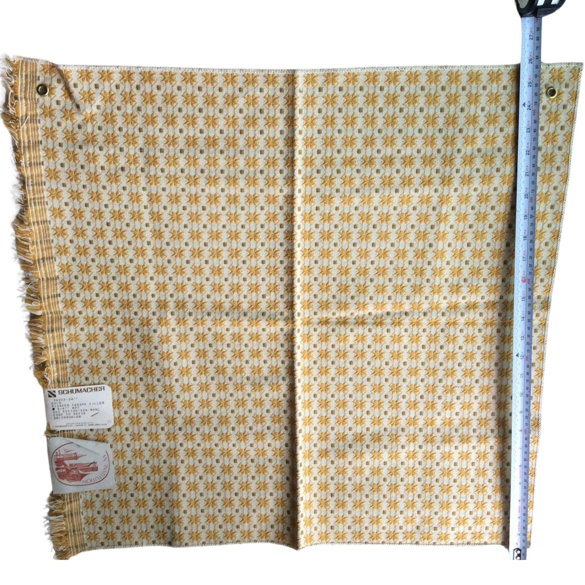 A square cream-colored woven textile with a repeating yellow and brown geometric pattern and fringed edges, shown flat with a blue tape measure along the right side and a product tag on the left.