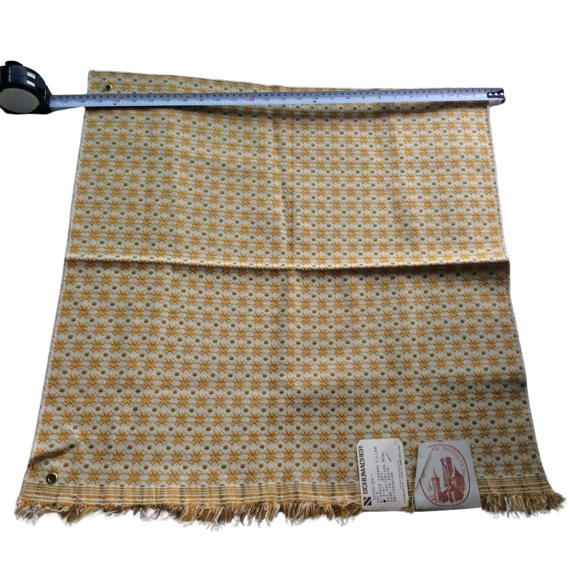1950s Schumacher wool fabric laid flat on a white background, featuring a small repeating geometric pattern in yellow, brown, and white. A metal tape measure is placed across the top edge to indicate size, and two tags are attached near the bottom fringe.