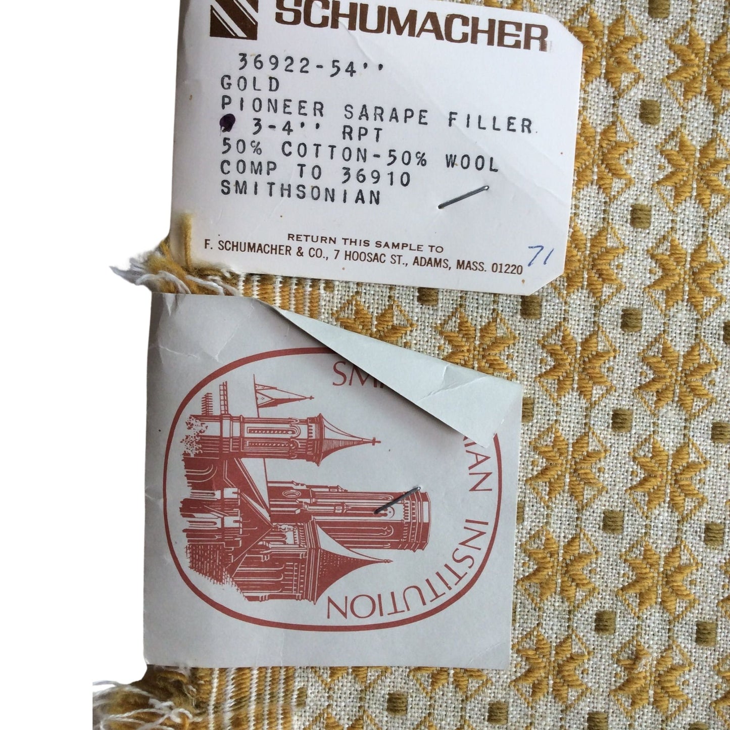 A close-up of a vintage patterned cotton-silk sarape fabric with gold and beige woven motifs, showing a white paper label with 'SCHUMACHER' and a red stamp from the Smithsonian Institution.