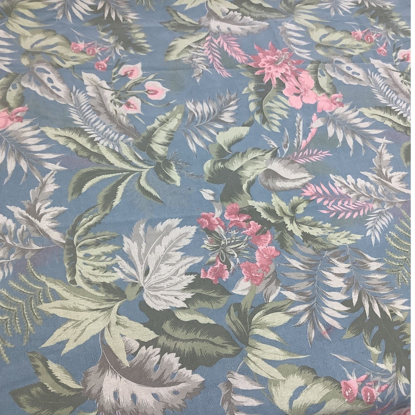 A close-up of a tablecloth featuring a tropical floral design with green, white, and pink leaves on a dusty blue background.