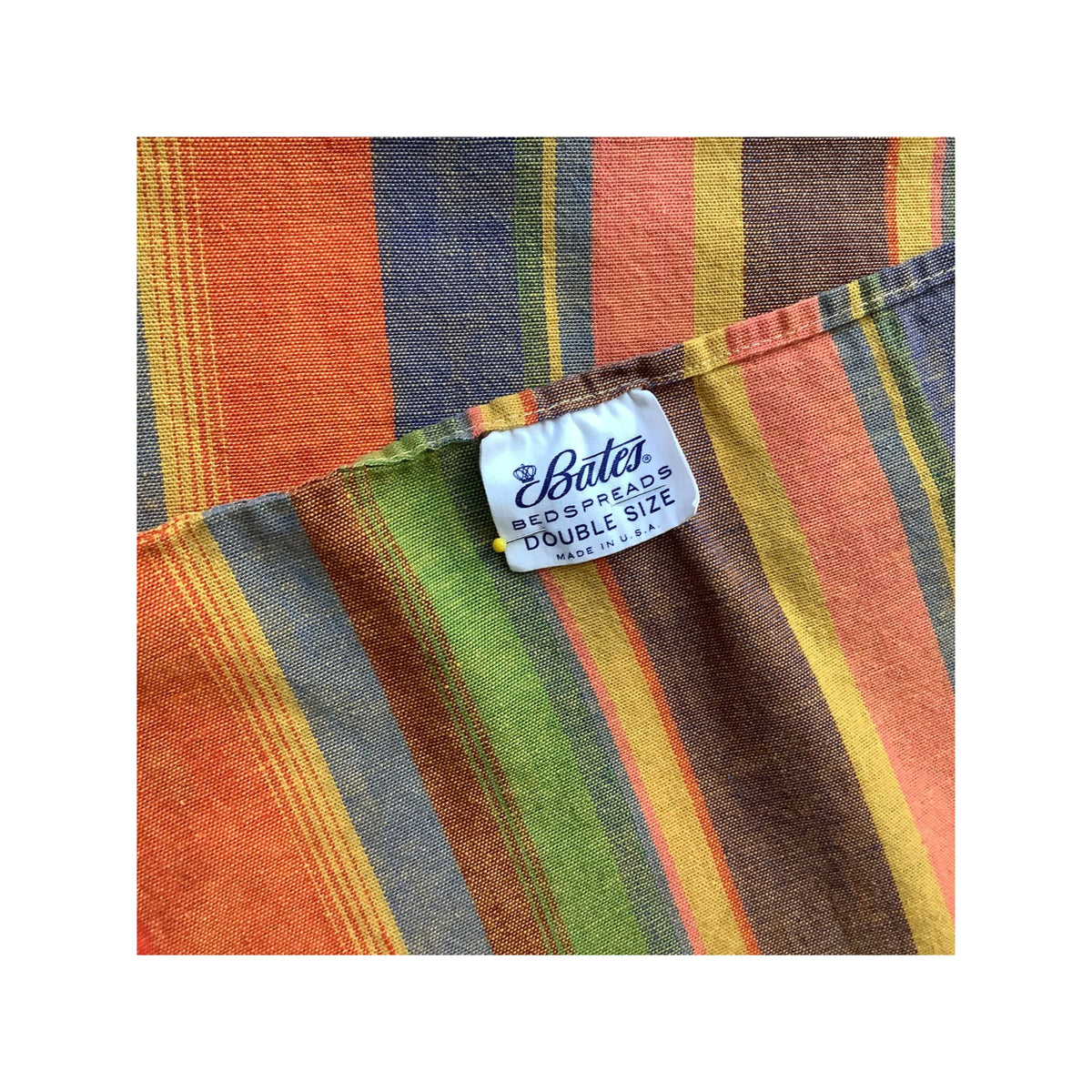 A close-up of a striped cotton bedspread with a visible white fabric tag reading 'Bates Bedspreads DOUBLE SIZE made in the U.S.A' The stripes feature orange, brown, green, yellow, and blue tones.