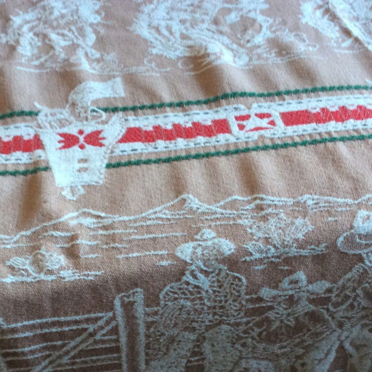 A close-up of a woven textile with a brown background, featuring white cowboys and landscape motifs, a red stripe with white stars, and green border lines.