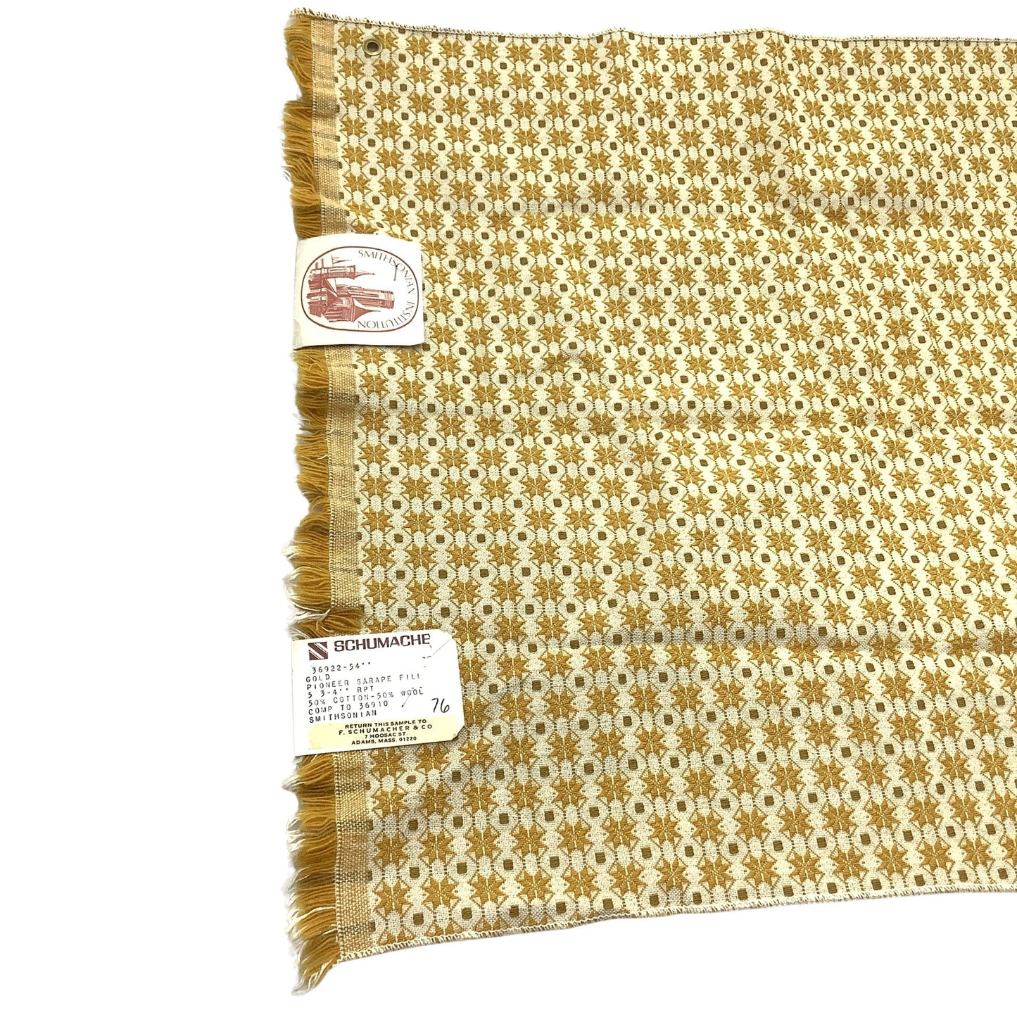 Vintage Schumacher fabric sample A square woven textile with fringe on one side, featuring a repeating geometric pattern in mustard yellow and off-white. Two paper tags are attached to the fabric.