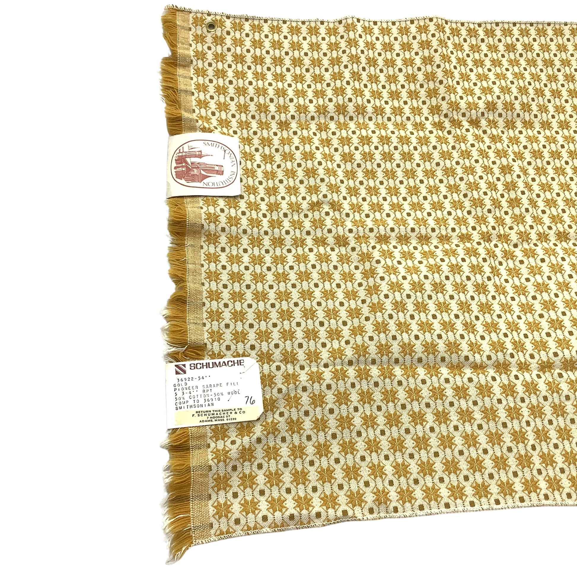 Vintage Schumacher fabric sample A square woven textile with fringe on one side, featuring a repeating geometric pattern in mustard yellow and off-white. Two paper tags are attached to the fabric.