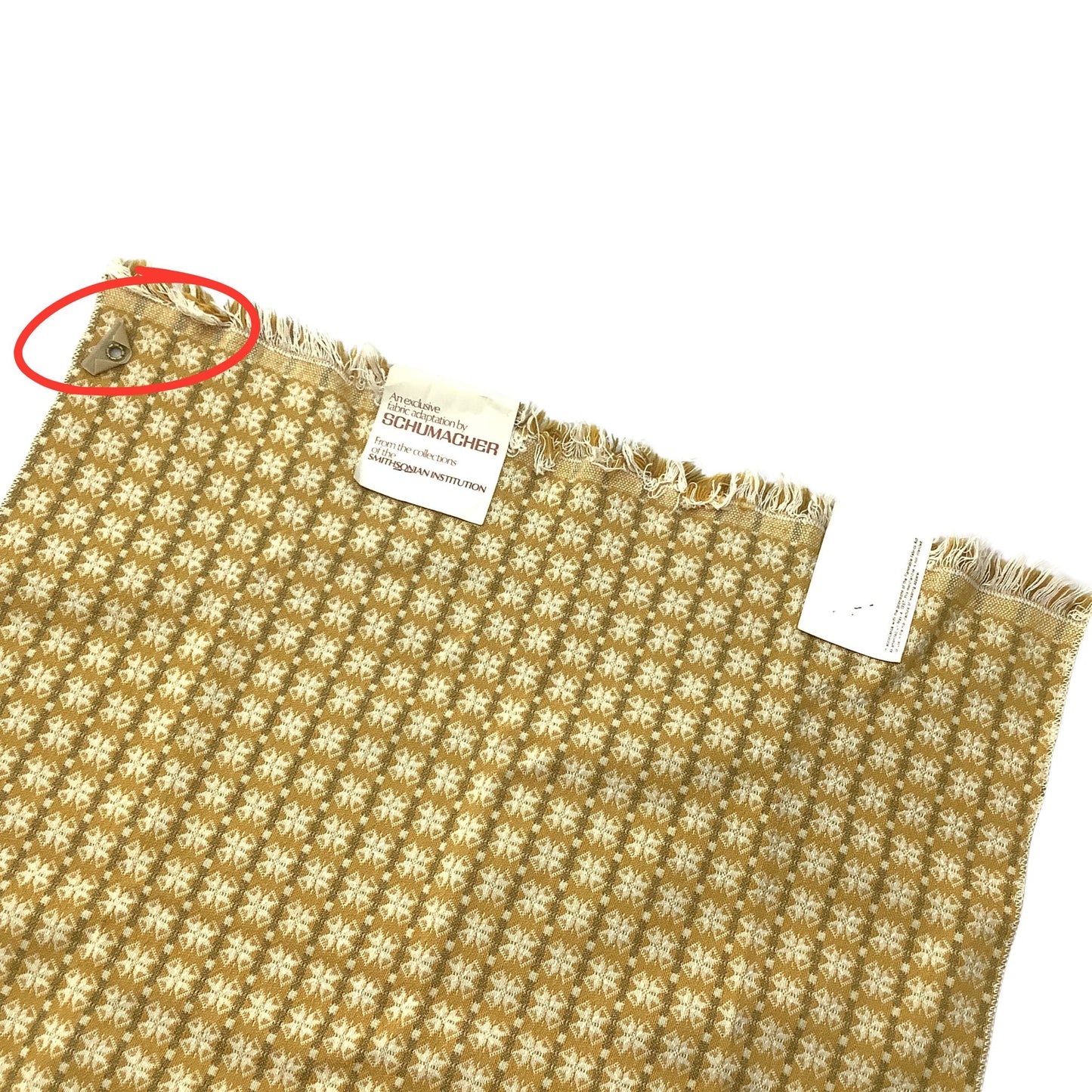 Vintage Schumacher fabric sample with a repeating white geometric patter on a mustard yellow background, featuring a frayed edge and a sewn-in label. A red circle highlights grommet at top left corner.