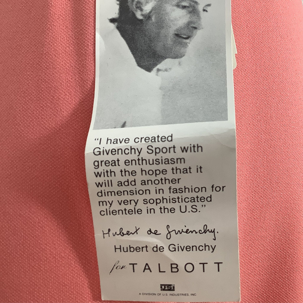 A vintage paper card or label with a black-and-white portrait at the top and a printed quote from Hubert de Givenchy, placed on a textured pink fabric background.