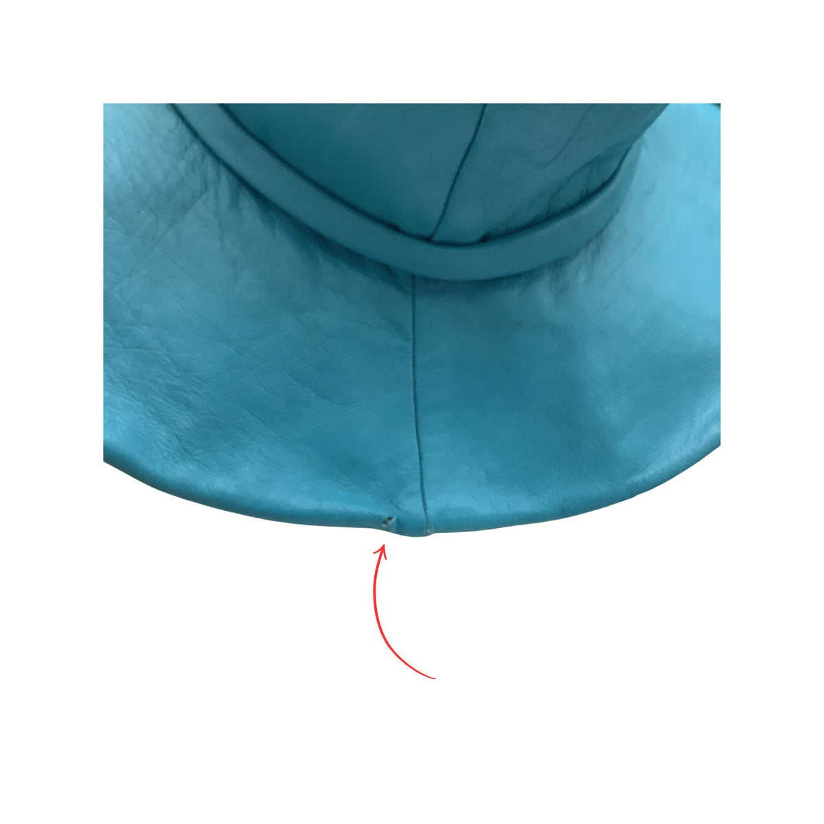 Close-up of the bottom edge of a teal blue hat or garment, showing a curved seam and a small red arrow pointing to small perforation area on a white background.