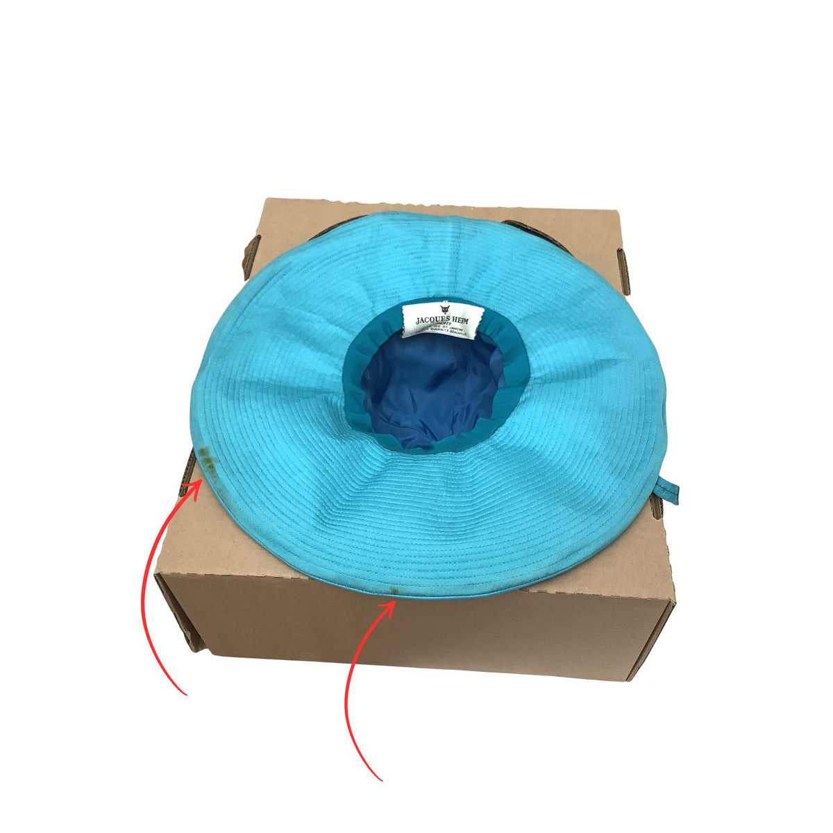 Jacques Heim blue leather hat resting on top of a brown cardboard box insert, photographed against a plain white background. Red arrows pointing to rust stains.