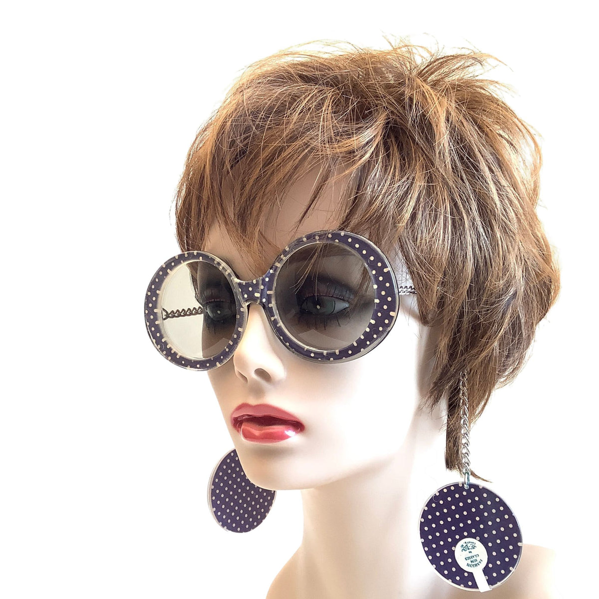 A mannequin head with a short brown wig wearing oversized round sunglasses with purpl polka-dot frames and matching dangling earrings, shown against a plain white background.