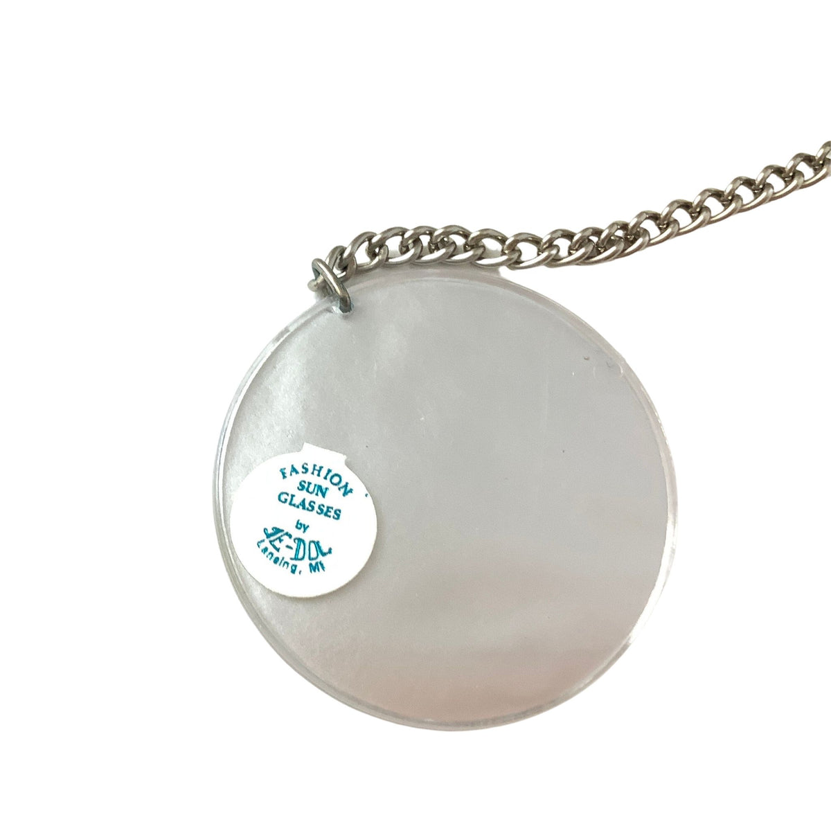 A round translucent silver-tinted fashion sunglasses disc earring on a silver-tone chain, with a white circular sticker reading 'FASHION SUN GLASSES by Je-Dol Lansing, MI' attached to the front.