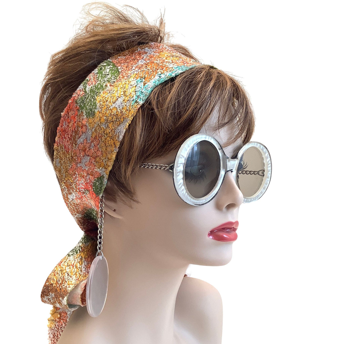A mannequin bust wearing a multicolor floral-patterned headscarf with dangling chain earrings and round silver-framed sunglasses, set against a plain white background.