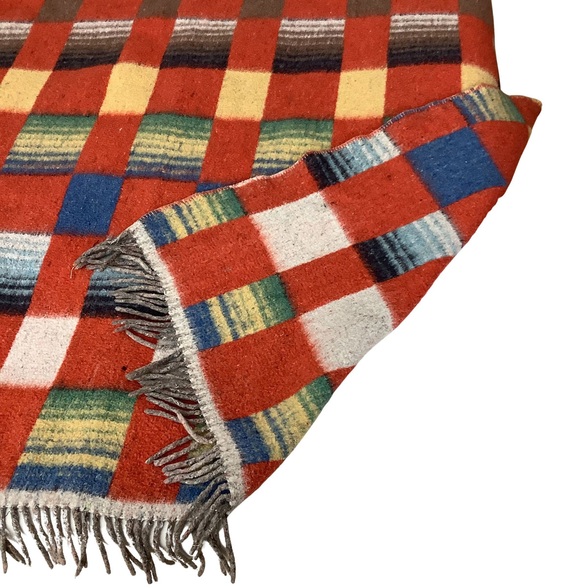 Folded corner of a wool blanket with a multicolor plaid pattern, featuring a dominant red base with blocks of white, blue, green, yellow, and dark brown, finished with gray fringe.