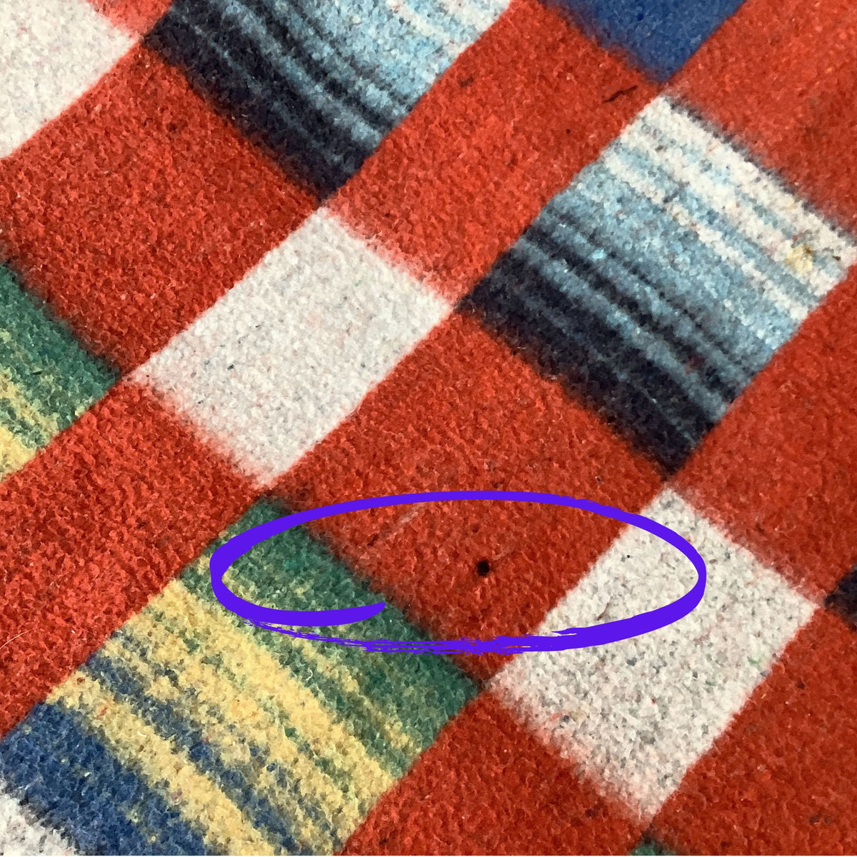 Close-up of a multicolor plaid fabric with red, white, blue, green, and yellow stripes, featuring a purple hand-drawn circle highlighting a small dark spot on the red section.