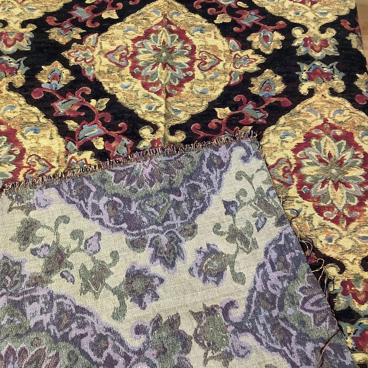 A close-up of a woven a textile showing two patterned sections: a black field with ornate gold, red, and green medallion motifs, and a light beige section with lavender, purple, and olive floral scrollwork. The textile fills almost the entire frame.
