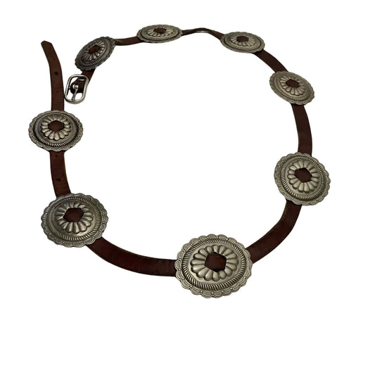 A western-style belt with a dark brown leather strap and eight large silver-toned conchos featuring dark brown centers, arranged in a circle on a white background.