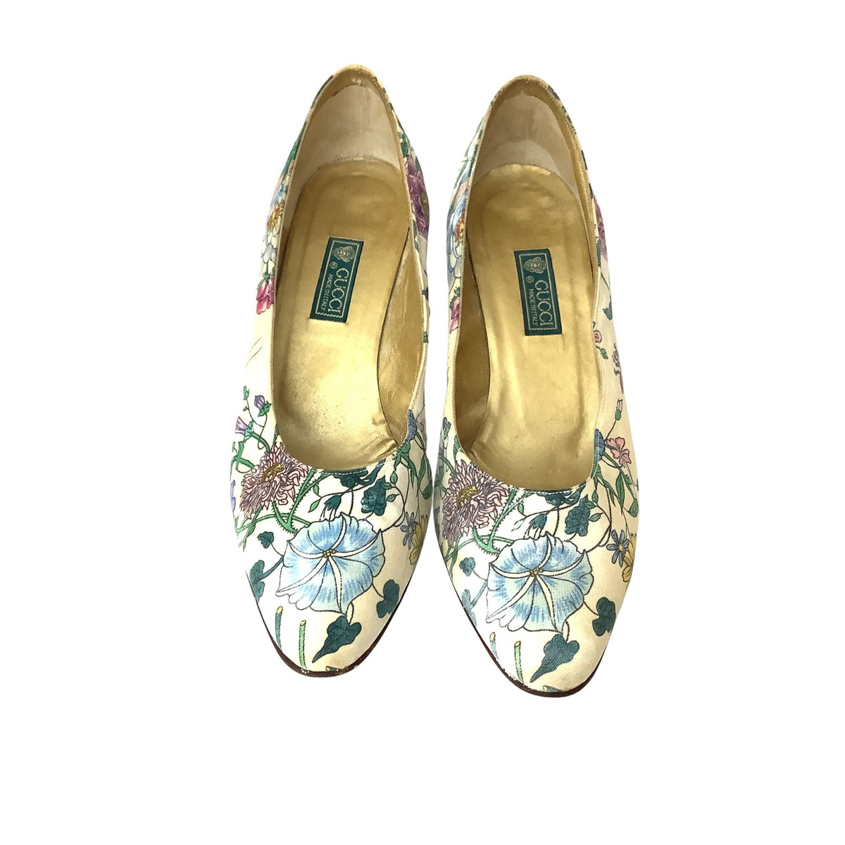 Vintage Gucci pumps with a cream-colored satin-like fabric covered in a detailed floral print featuring blue, purple, and green flowers and leaves. The interior of the shoes is lined with a gold-colored material, and a green brand label reading 'Gucci' is visible on each shoe.