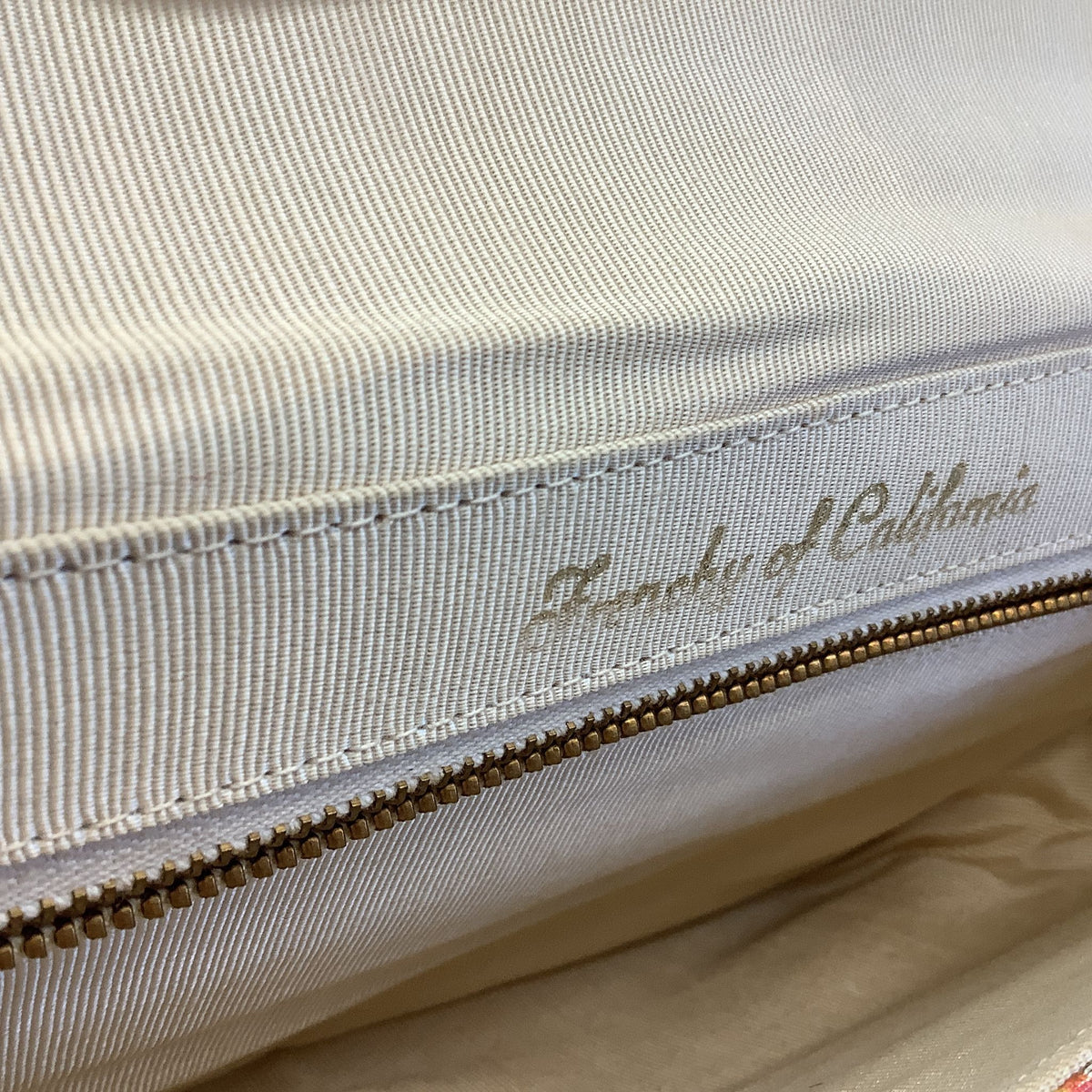 Purse Interior close-up of a cream-colored fabric zipper with a white tape pull tab featuring the script logo 'Frenchy of California' in gold, set against a matching cream fabric background.
