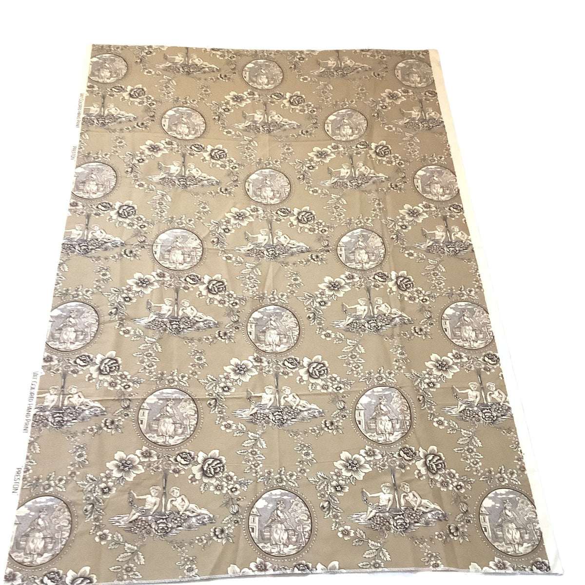 A rectangular piece of fabric or wallpaper with a repeating toile-style pattern, laid flat against a white background. The design features circular medallions with figures, floral motifs, and basket scenes in muted gray and taupe tones over brown base.