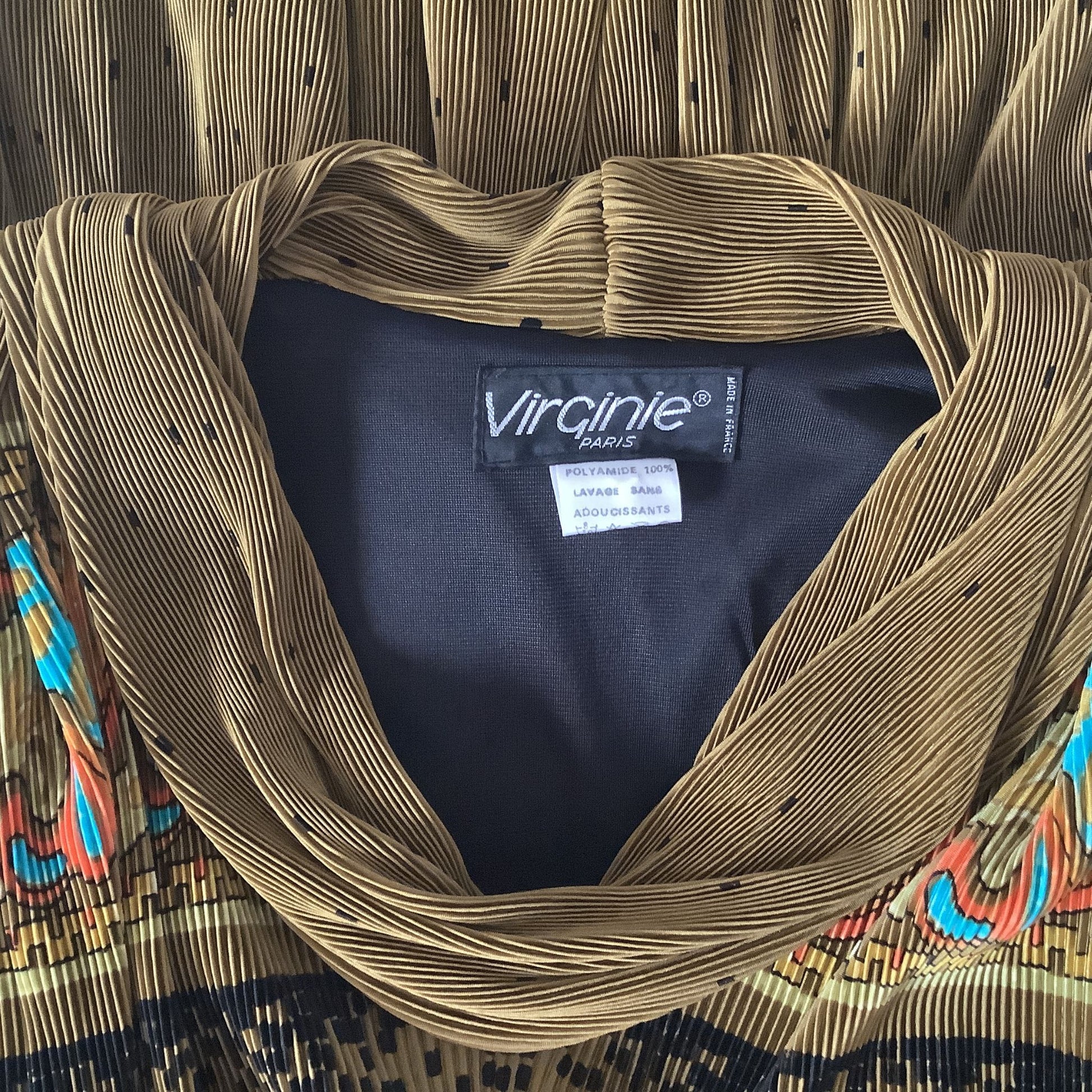 A close-up of a pleated top with a V-neckline, featuring a gold and black pleated fabric with a colorful patterned border. The inside collar shows a brand label for 'Virginie PARIS'.