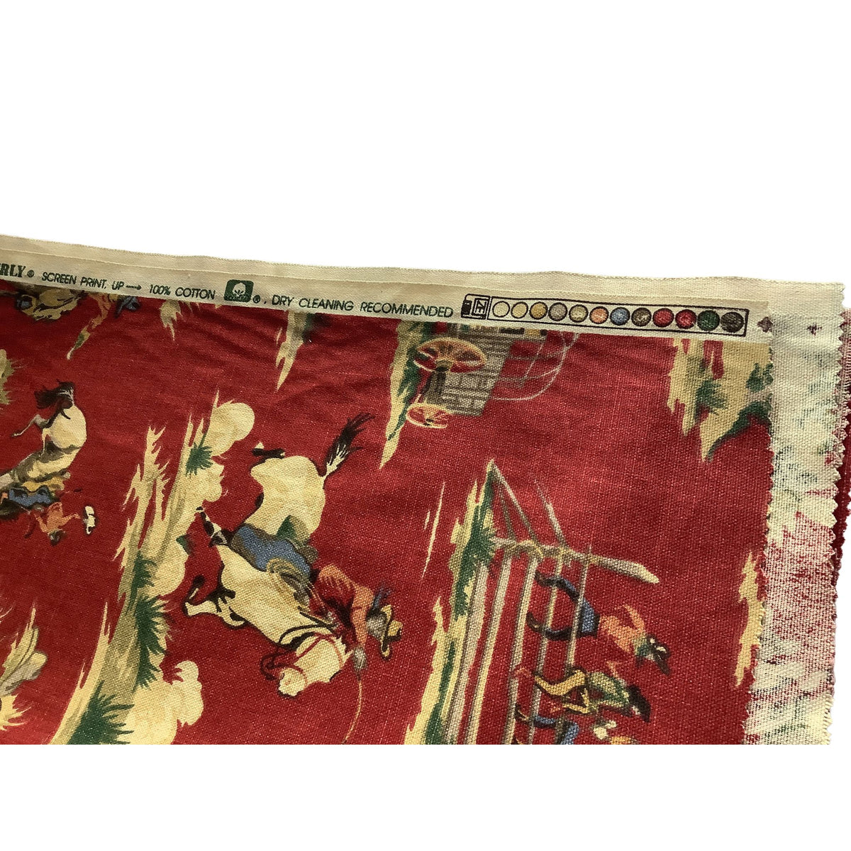 A close-up of a vintage red cotton screen print fabric featuring a detailed folkloric scene of figures on horseback and a cowboy, with a cream-colored selvage edge showing cleaning tag printed text.