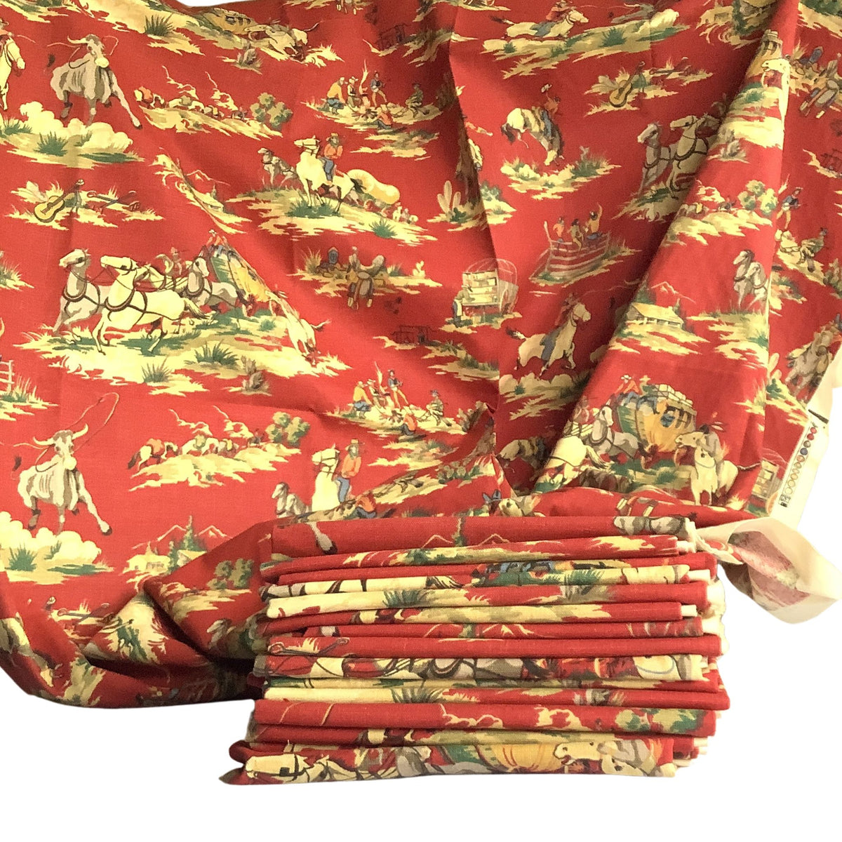 A folded stack of southwestern red fabric with a vintage cowboy and cattle scene toile pattern, displayed next to a larger matching fabric roll on a white background.